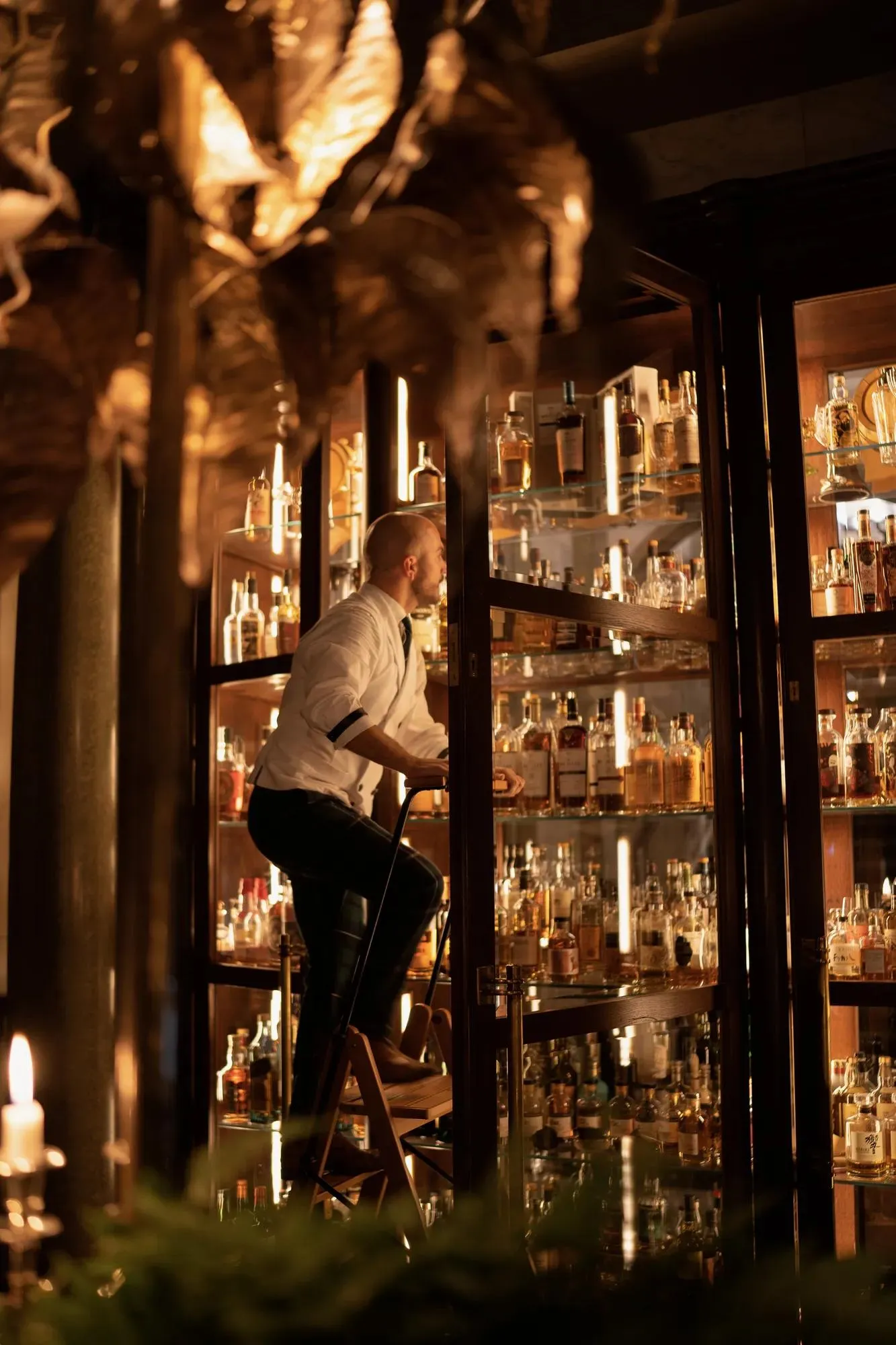 A bartender reaching for bottles behind the warmly lit bar at Scarfes Bar